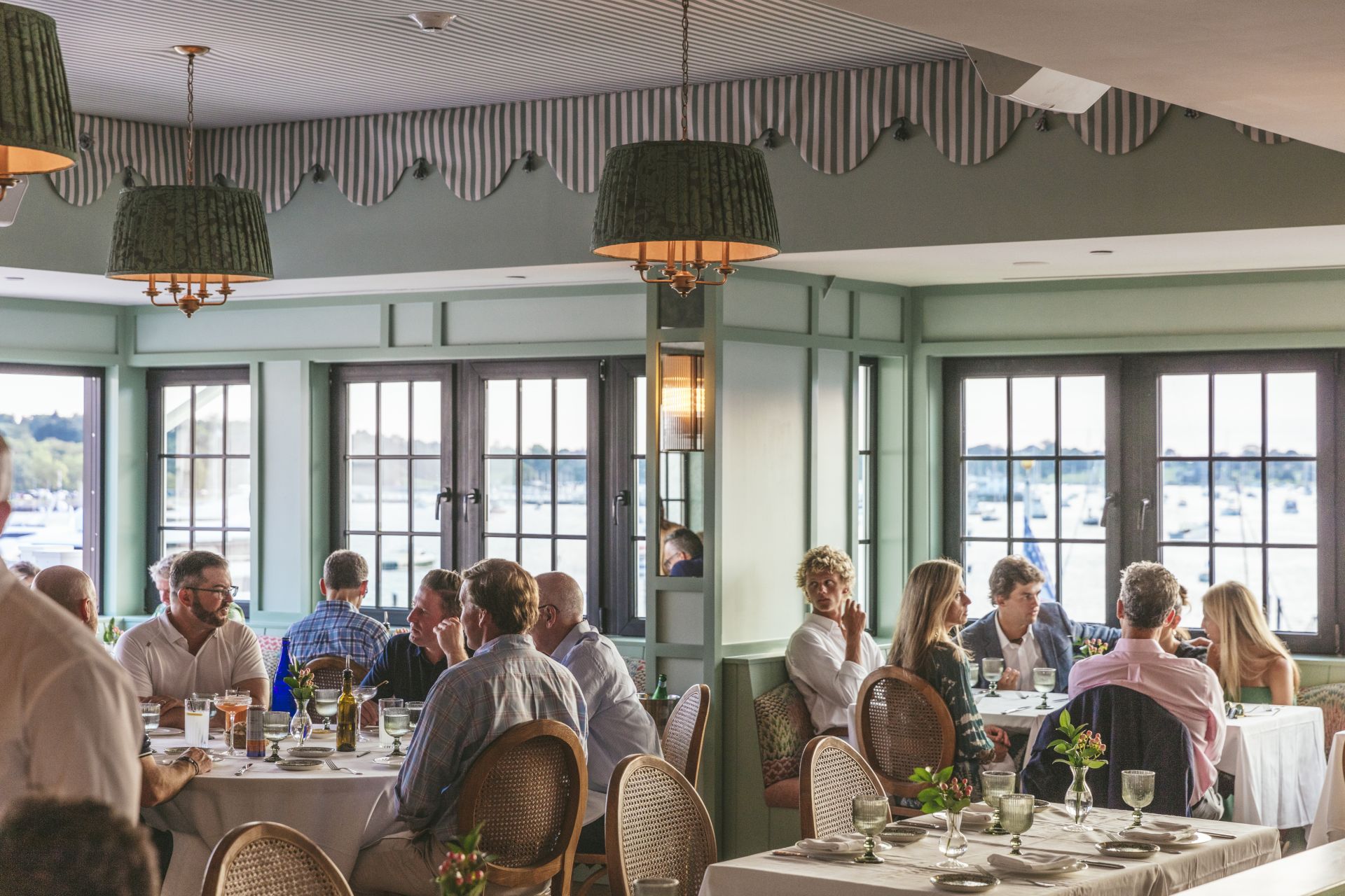 People dining in a restaurant with large windows and a view of boats outside. The room has green accents and decorative hanging lights.