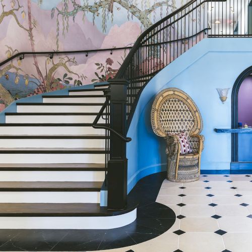 A stylish interior with a staircase, colorful mural, wicker chair, and elegant floor tiles, adjacent to an arched reception counter.