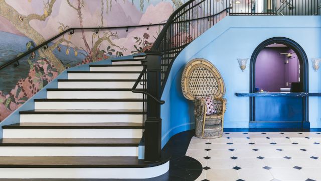 A curved staircase with a black railing and white steps leads up to a mural-covered wall, with a wicker chair and archway to the right.