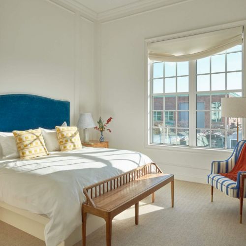 A bright bedroom with a large bed, striped chair, and wooden bench. The room features a big window with natural light.