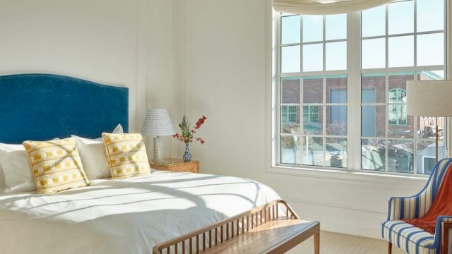 A neatly made bed with white bedding and yellow pillows, a wooden bench, and a striped chair in front of a large window.