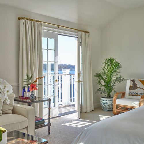 A bright room with a bed, white curtains, a chair, a small table with flowers, and a door opening to a balcony with a railing.