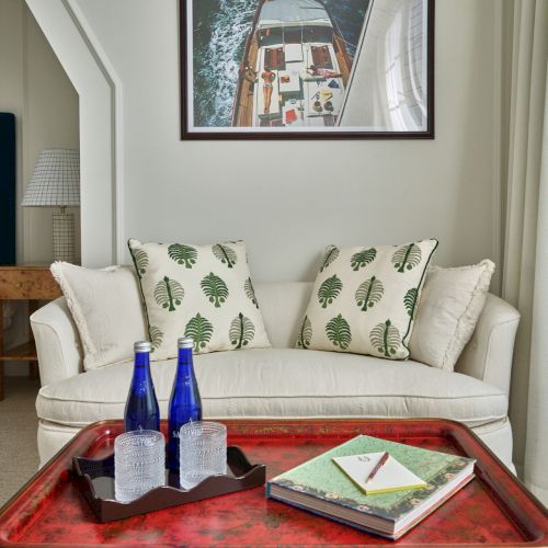 A cozy living area with a white sofa, leaf-patterned pillows, and a red table featuring books and blue bottles. A yacht photo is on the wall.