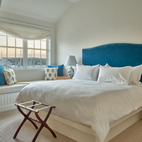 A cozy bedroom with a large bed, white bedding, blue headboard, window seat, and a view outside with soft lighting from lamps on both sides.