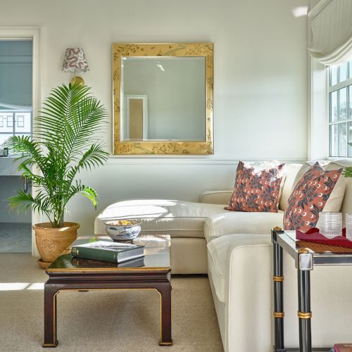 A cozy living room with a white sectional sofa, colorful cushions, a potted plant, books, a mirror, and a coffee table with decor.