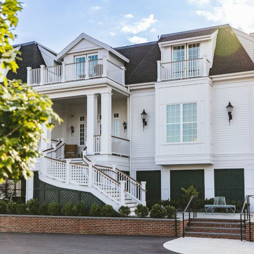 A large, elegant two-story house with white siding, multiple balconies, and a staircase leading to the entrance, surrounded by trees.