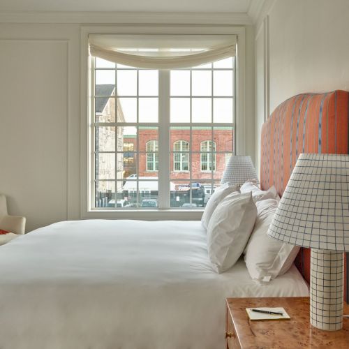 A cozy bedroom with a large bed, grid-patterned lamps, and a window view of buildings. There's a chair with an orange blanket by the wall.