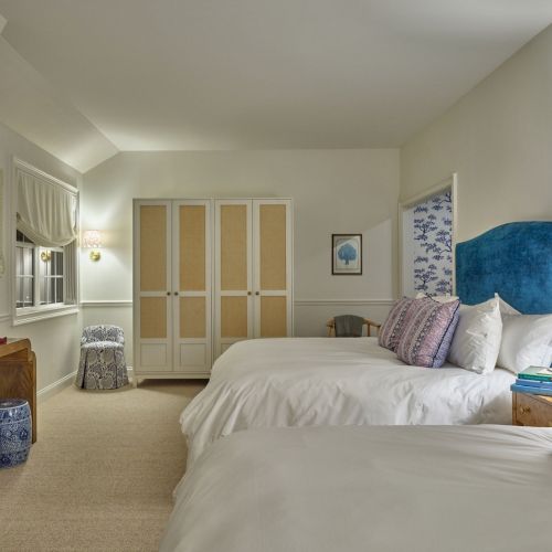 A cozy bedroom with two beds, blue headboards, a wooden desk, wicker baskets, and decorative items, creating a calm atmosphere.