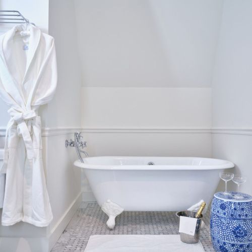 A white bathroom features a clawfoot tub, bathrobe, blue stool, towel, and decorative items, creating a serene and stylish space.