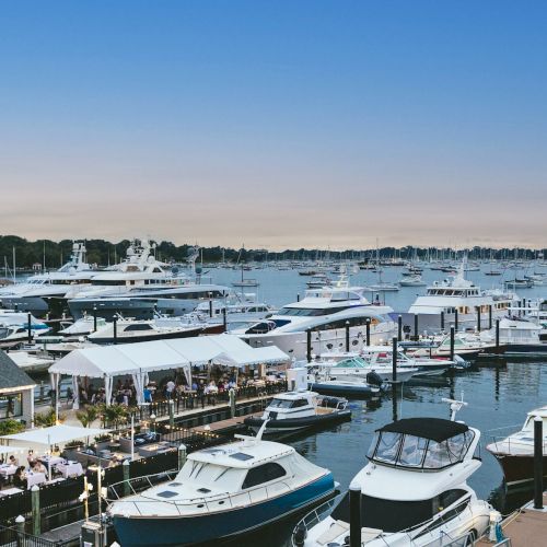 The image shows a marina with numerous boats and yachts docked, alongside a waterfront dining area under a clear evening sky.