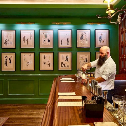 A bartender works at a wooden bar with framed art on green walls. Various bottles and tools are visible on the counter.