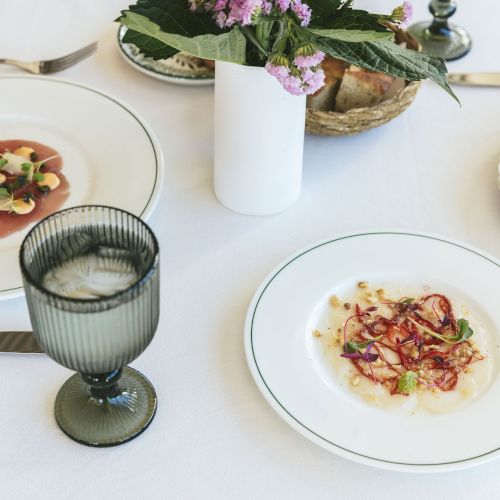 A table with three dishes of elegant food, a green glass with ice, a vase with flowers, and a basket of bread on a white tablecloth.