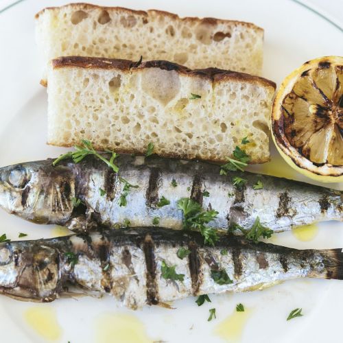 Grilled sardines with herbs, slices of bread, and a half lemon are on a white plate.