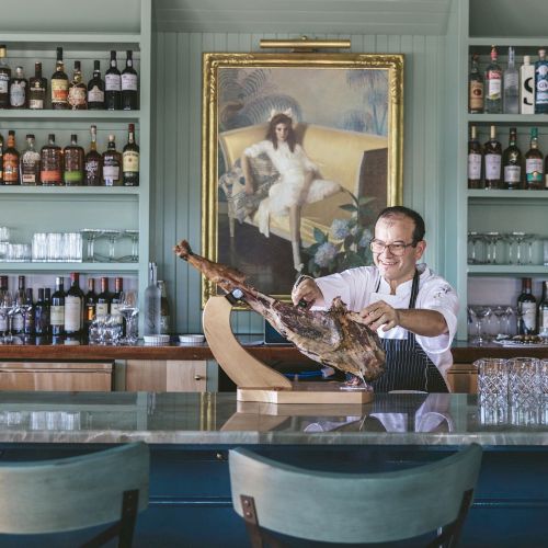 A man in a bar is slicing a large meat leg, with shelves of bottles and a dog portrait behind him. The bar has teal chairs in front.