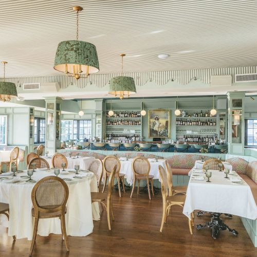 The image shows an elegant restaurant interior with round tables, chairs, soft lighting, and a bar in the background, creating a cozy ambiance.