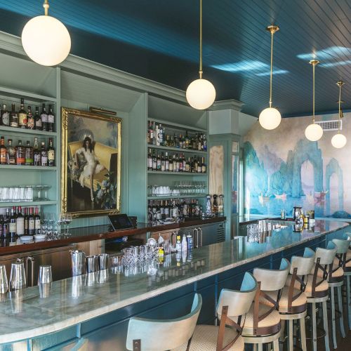 A stylish bar with a marble countertop, shelves of bottles, hanging globe lights, mural, painting, and rows of stools.