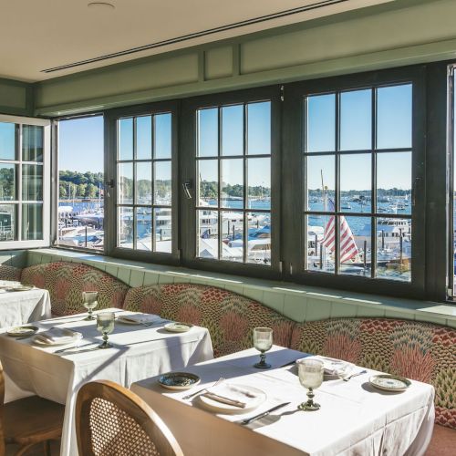 Cozy restaurant interior with tables by a window overlooking a marina with boats, calm water, and clear skies outside.