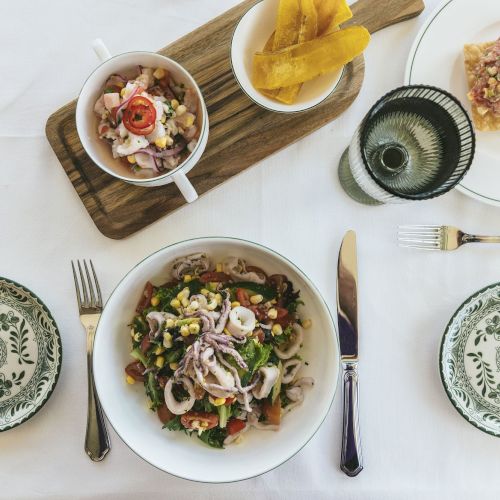 A dining setup with salad, seafood, and plantain chips on a wooden board, accompanied by utensils and patterned bowls on a white tablecloth.