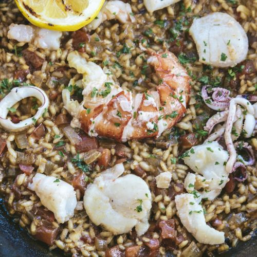 A seafood rice dish in a pan with shrimp, scallops, squid, herbs, and a lemon slice on top.