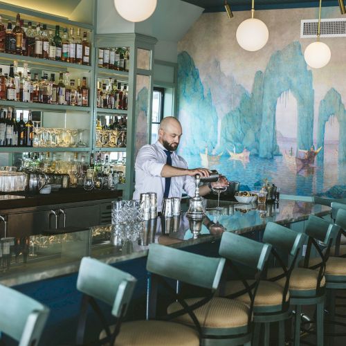 A bartender is preparing drinks at a stylish bar with a mural of scenic landscapes, surrounded by shelves of bottles and elegant bar stools.