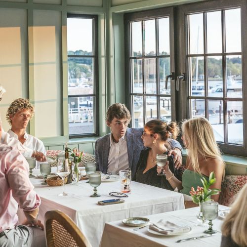 A group of people sitting at a dining table in a restaurant with large windows, looking out at a marina or waterfront.