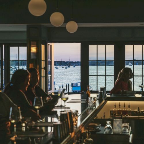 A dimly lit bar with people enjoying drinks, overlooking a waterfront with boats and a sunset in the background.