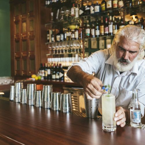 A bartender is preparing a cocktail at a bar with a variety of bottles on shelves in the background, focusing intently on his work.