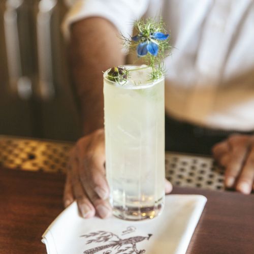 A person serves a tall, light-colored cocktail garnished with a flower, resting on a folded napkin.