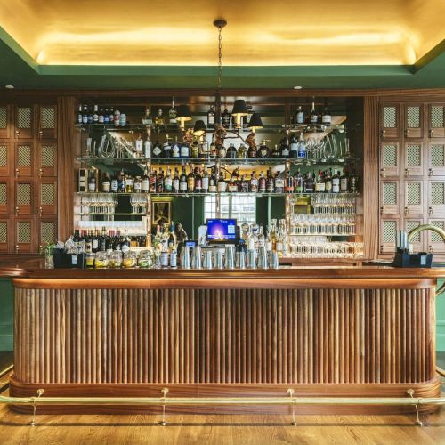 A stylish bar with a wooden counter, bottles lined up on shelves, green walls, and artwork. The interior looks elegant and organized.