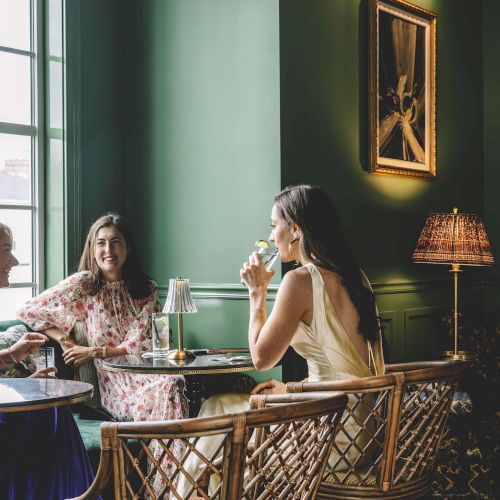 Three women are sitting at a table in a stylish, green-painted room, enjoying drinks and chatting by a window with natural light.