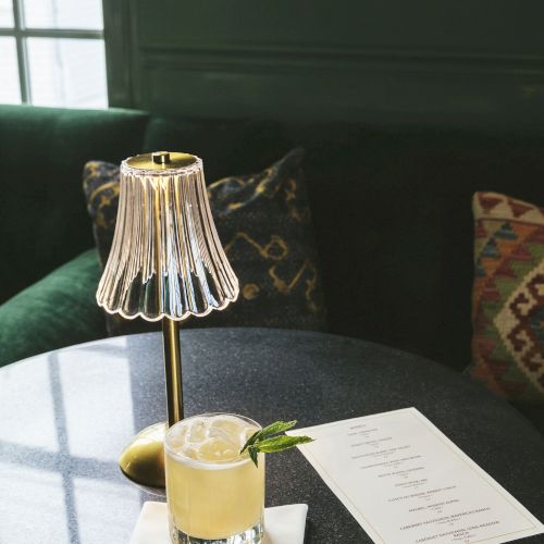 A cozy setting with a drink on a table, a stylish lamp, and a menu. The ambiance is intimate and inviting.