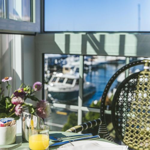 A table set for breakfast with orange juice, plates, and flowers overlooks a harbor with boats under a clear blue sky.