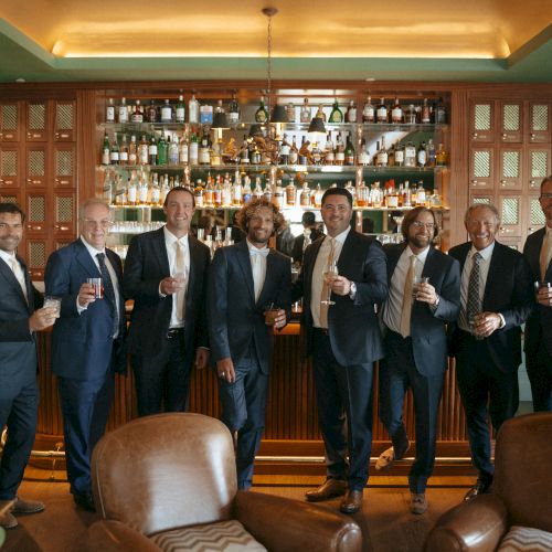 A group of eight men in suits stand in front of a bar holding drinks, with leather chairs in the foreground, in a warmly lit room.