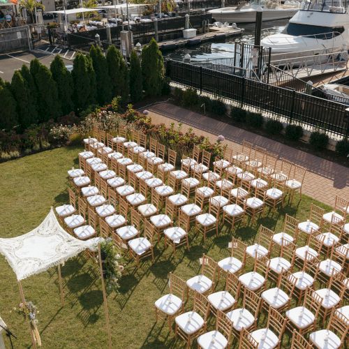 Outdoor wedding setup with rows of chairs facing a canopy, situated by a marina with yachts in the background.