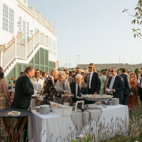 A group of people is gathered at an outdoor event, with a buffet table set up and a building in the background.