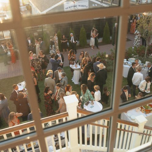 Aerial view of a formal outdoor gathering with people mingling around tables, seen through a window.