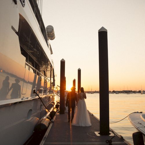 A bride and groom walk along a dock beside a yacht with a sunset in the background, creating a romantic scene beside the water.