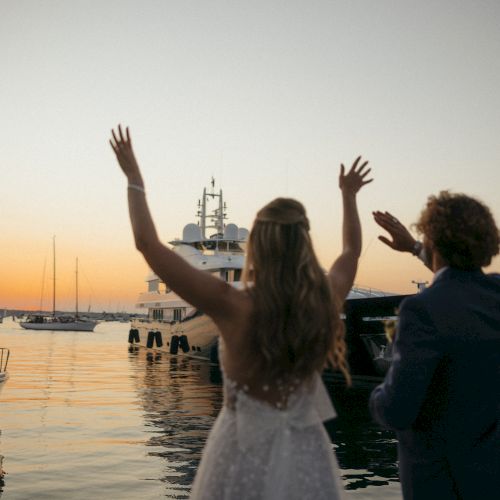 A couple waves at a yacht during sunset on a dock.