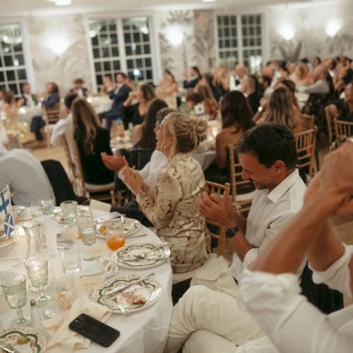 A large group of people seated at tables in a fancy event room, clapping and watching someone speak at the front.