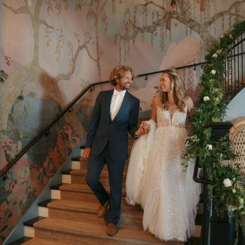 A couple in wedding attire descends a decorated staircase with floral arrangements and mural backgrounds.