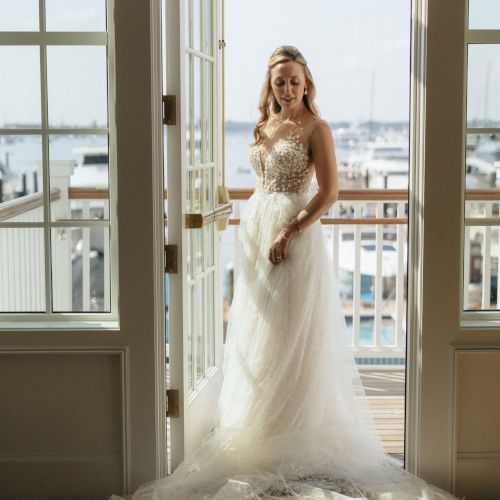 A person in a wedding dress stands in a sunlit doorway, looking outside towards a marina.