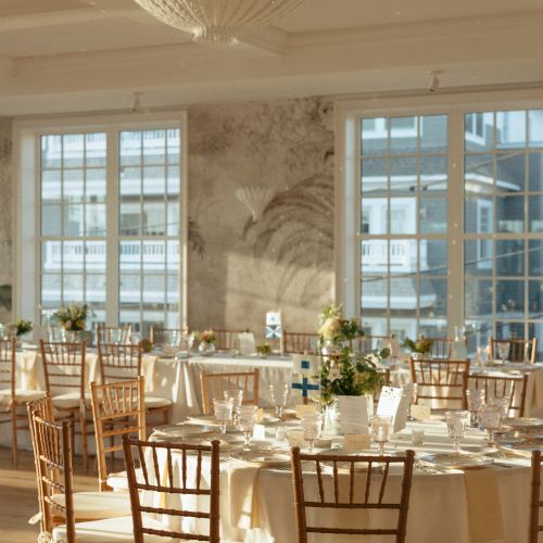 A sunlit banquet room with round tables, elegant wooden chairs, white tablecloths, and floral centerpieces, featuring large windows.