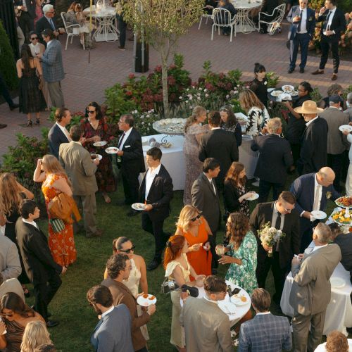 A large outdoor gathering with people in formal attire socializing, eating, and drinking around tables on a patio.