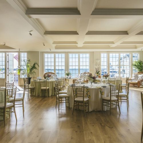 A bright, elegant event space with round tables, chairs, and large windows overlooking water, decorated with plants and floral arrangements.