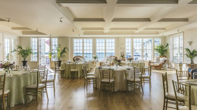 A brightly lit dining room with round tables set for an event, featuring white chairs, green tablecloths, and floral arrangements.