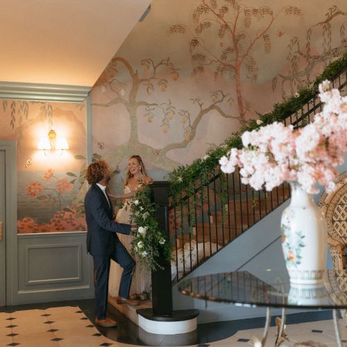 A couple stands on a staircase in an elegant room with floral wallpaper, a chandelier, and floral arrangements, creating a romantic setting.