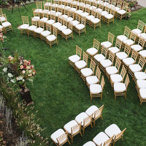 An outdoor wedding setup with rows of white-cushioned chairs arranged on grass, surrounded by floral decorations and greenery.