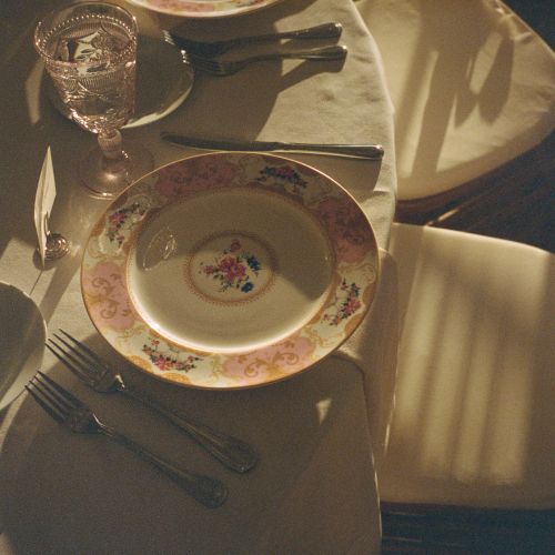 A table set with elegant floral plates, forks, knives, a glass, and a cloth napkin on a sunlit, round table.