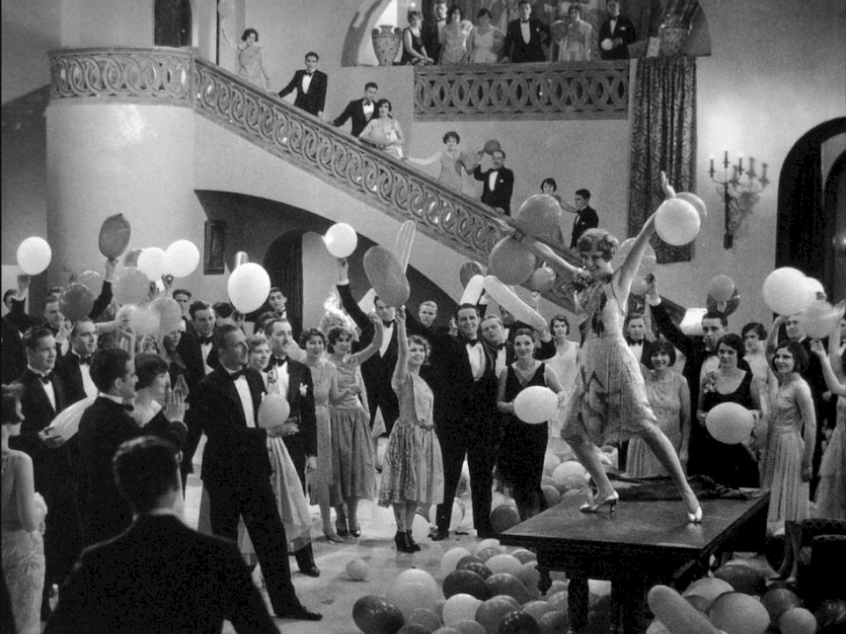A black-and-white party scene with people dancing and balloons, a grand staircase, and guests mingling around a central performer on a small platform.