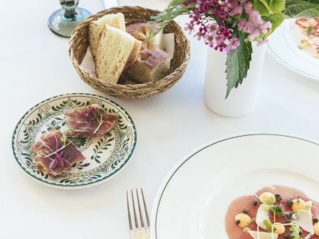 A fancy table setting with a bread basket, decorative plate, a vase of pink flowers, and a plated dish with pink sauce and toppings, plus cutlery and a plate.